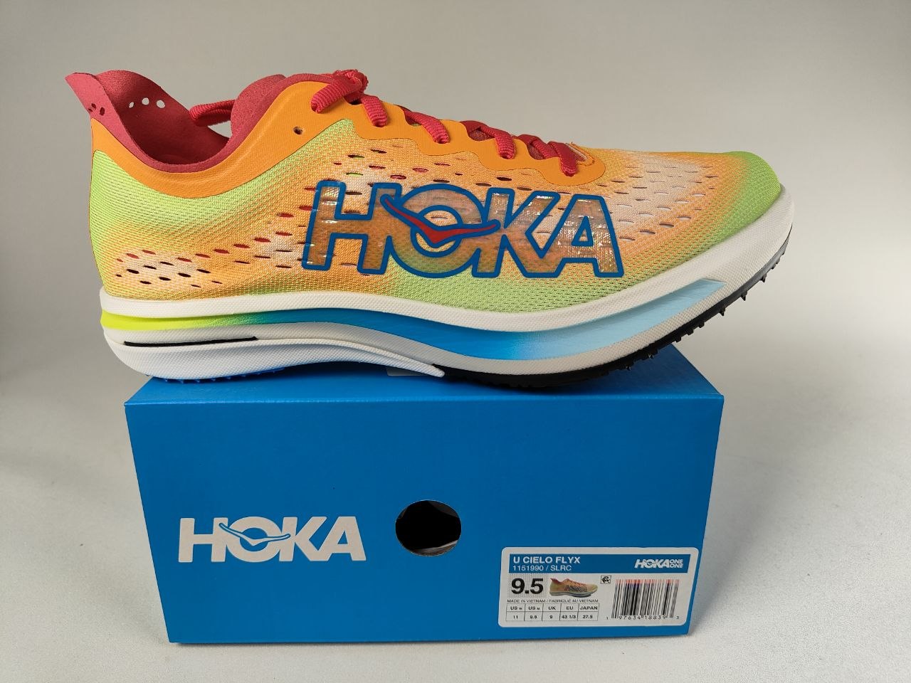 A Hoka Cielo FlyX Carbon-Plated Track Spikes Solar Flare with red laces on a blue box with a white Hoka branding on a light gray background – detail View @RunInRain
