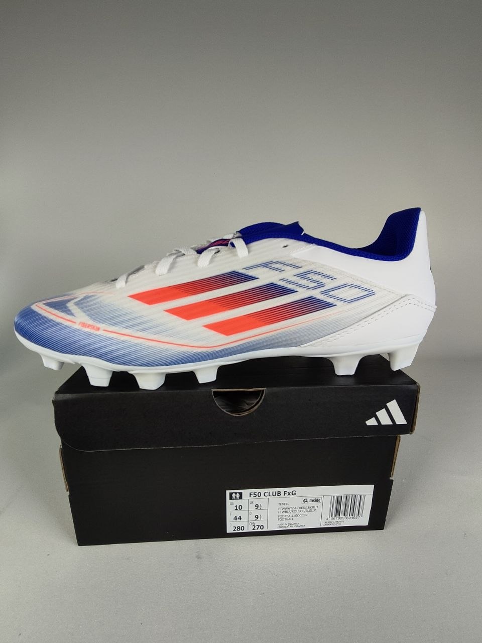 White Adidas F50 Club FxG with blue and red accents on a black Adidas box against a gray background