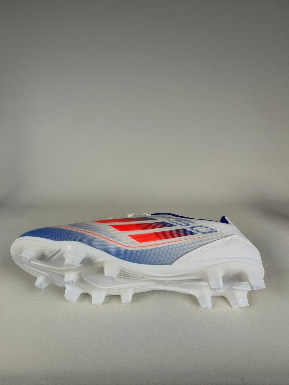 White Adidas F50 Club FxG cleat with red and blue design on a gray background - Side & Sole view @runinrain