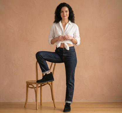 Woman in a white shirt and dark jeans sitting on a wooden stool against a beige wall, wearing Black Clarks Originals Desert Boot.
