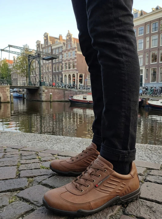 Person wearing brown shoes on a cobblestone street with canal and buildings in the background
