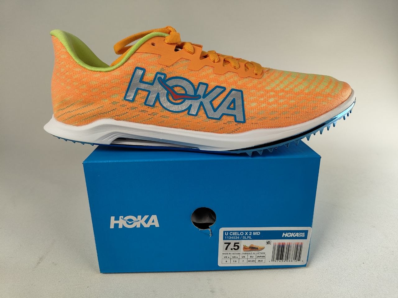 Orange HOKA Cielo X 2 MD track spikes with 'Hoka' branding on a blue shoe box - Side View