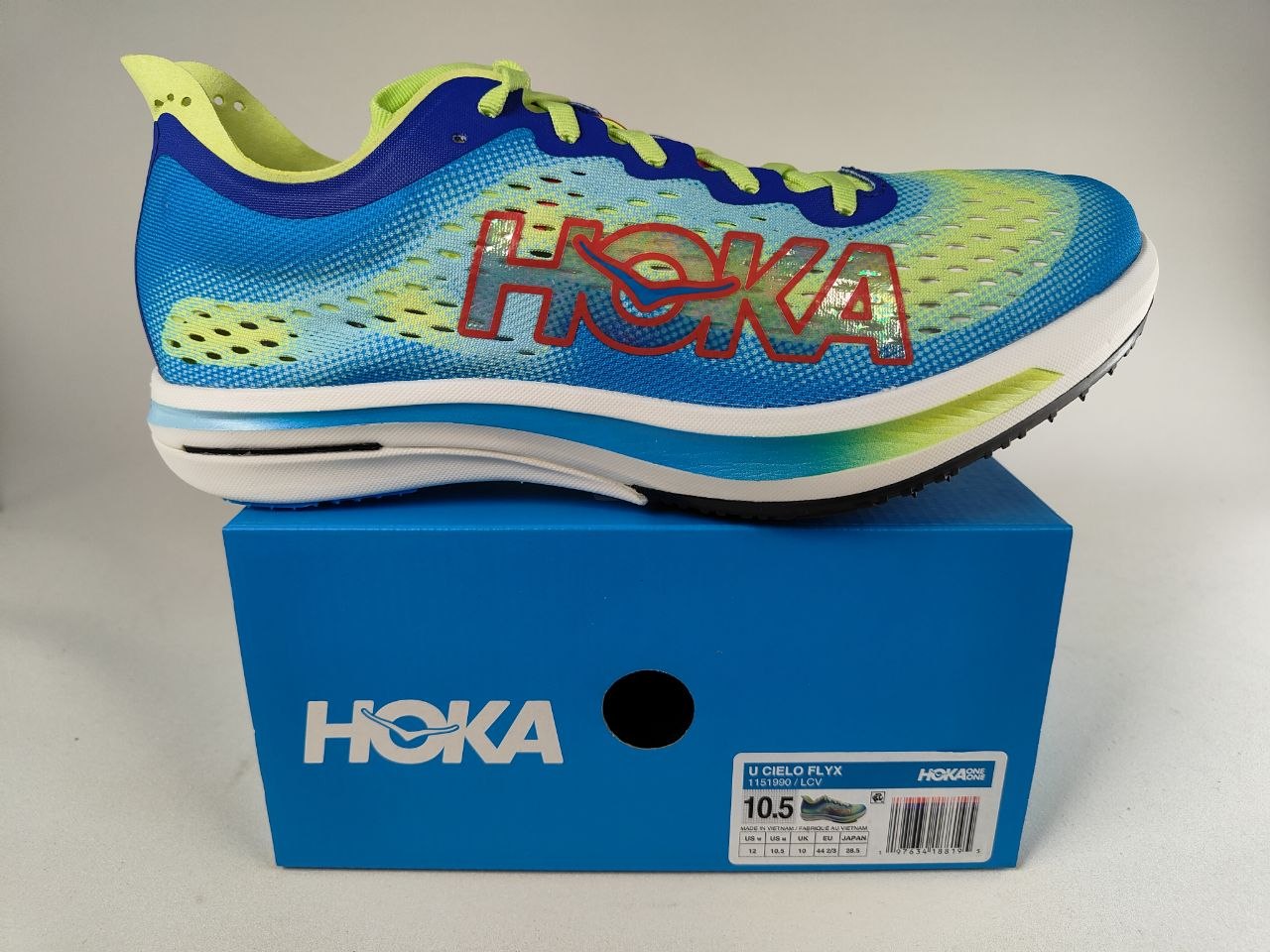 A pair of Hoka Cielo FlyX Carbon-Plated Track Spikes Cyan/Lime shoes with 'Hoka' branding on a Hoka-branded box on a white background – Detail View @RunInRain