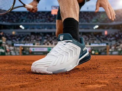 Tennis player's foot wearing a white and blue K-Swiss Ultrashot 4 Clay Men's Tennis Shoe on a clay court – free style @RunInRain