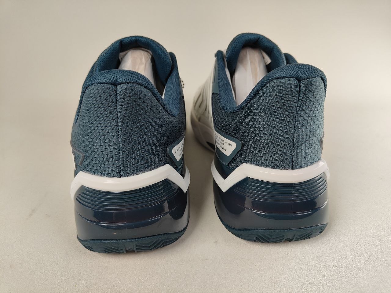 A pair of K-Swiss Ultrashot 4 men's clay court tennis shoes in white and dark blue on a beige background - Rear view @RunInRainwith.